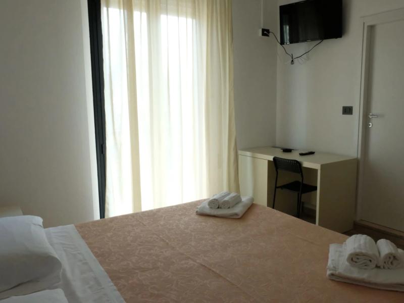 Camera semplice con letto matrimoniale, scrivania, TV e tende chiare.