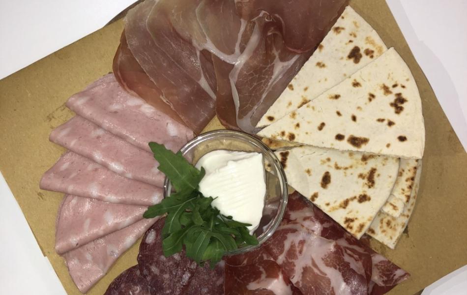 Tagliere con salumi, formaggio, rucola e piadina su carta marrone.